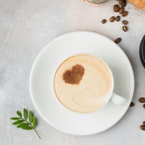 A cappuccino with a heart-shaped cocoa dusting on a saucer, surrounded by coffee beans.