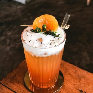 A vibrant citrus cocktail garnished with thyme and peach slice on a wooden table.
