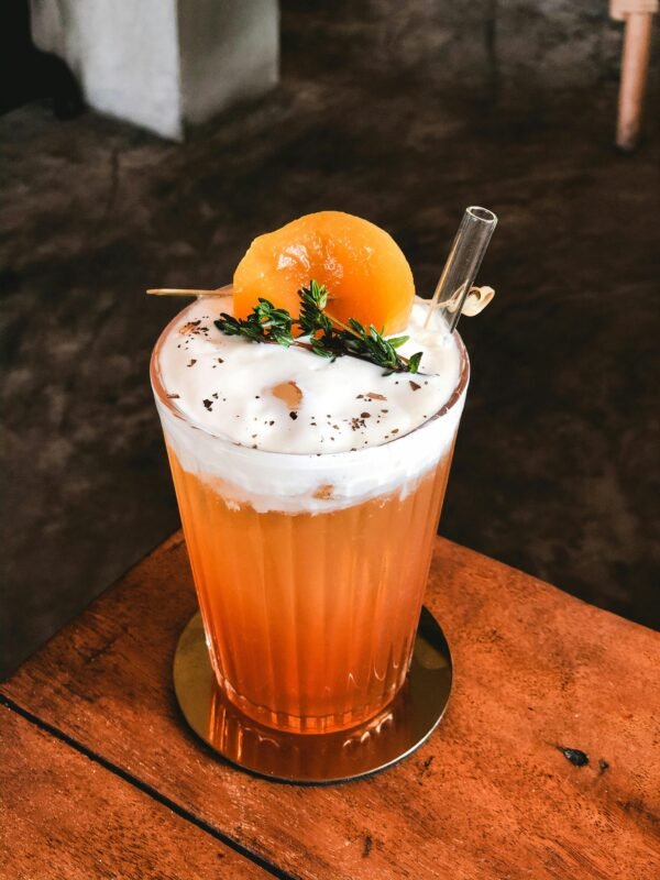A vibrant citrus cocktail garnished with thyme and peach slice on a wooden table.